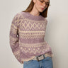A person wearing the Sanctuary Clothing fairisle crew neck sweater in twilight mauve multi stands against a plain off-white background, paired with tinted sunglasses, light blue jeans, and a gray belt.