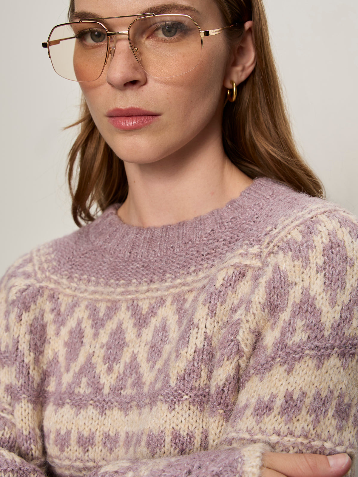 A woman with light brown hair wears gold-rimmed glasses, gold hoop earrings, and the Sanctuary Clothing fairisle crew neck sweater in twilight mauve multi. She gazes slightly to the side with a neutral expression.