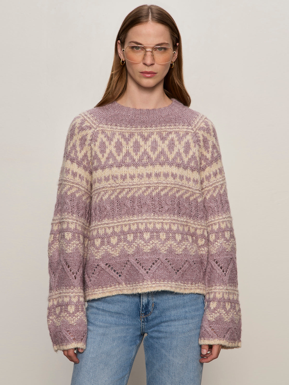 A person with straight, light brown hair wears clear glasses, the Sanctuary Clothing fairisle crew neck sweater in twilight mauve multi, and light blue jeans, standing against a plain light background.