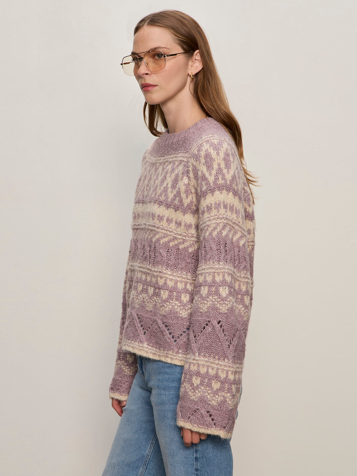 A woman with straight, light brown hair and glasses models the Sanctuary Clothing fairisle crew neck sweater in twilight mauve multi with blue jeans, standing sideways against an off-white background.