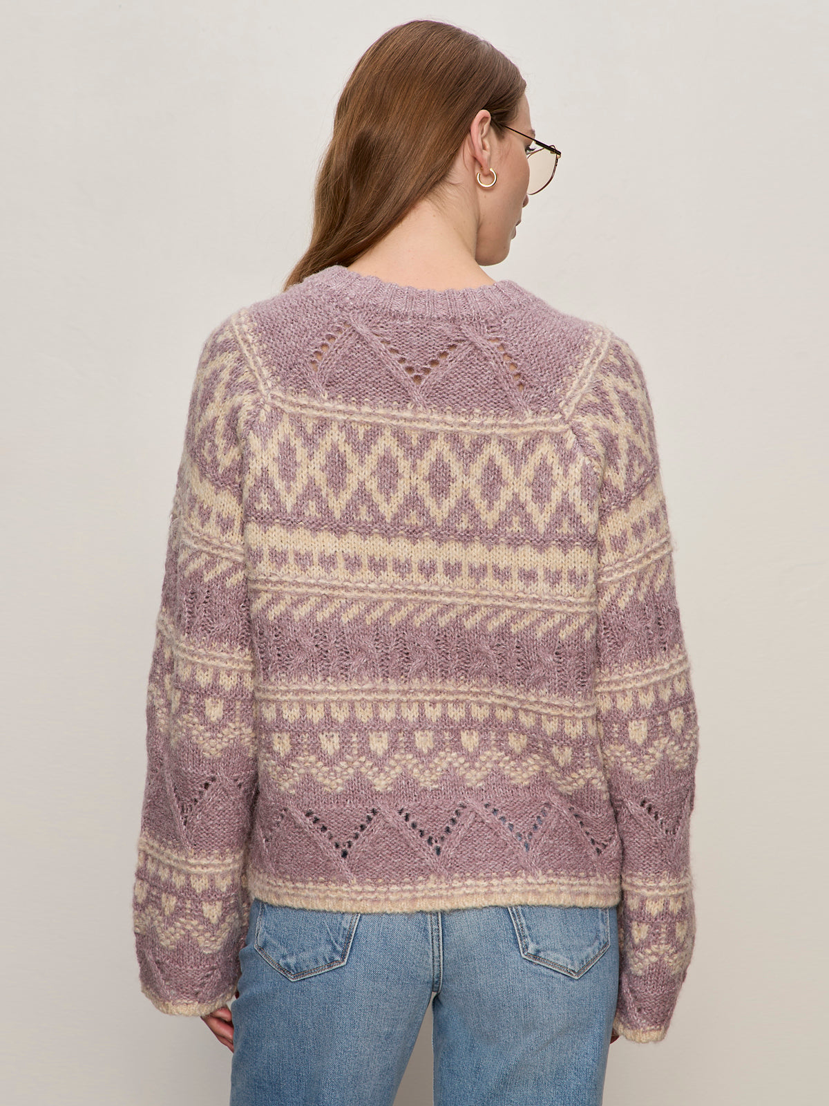 A woman with long brown hair and glasses wears the Sanctuary Clothing fairisle crew neck sweater in twilight mauve multi, paired with light blue jeans, facing away from the camera against a plain light background.