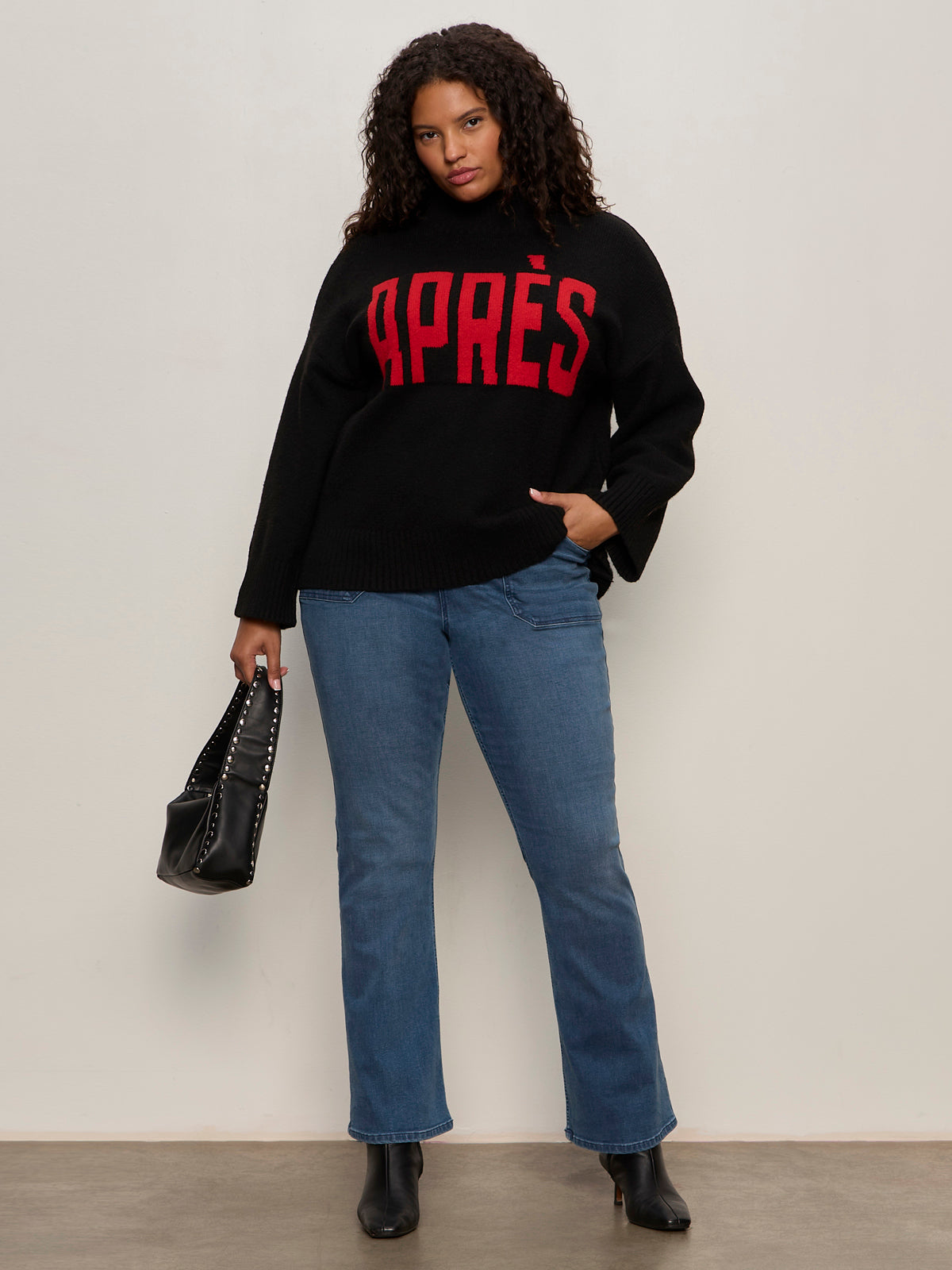 A woman with curly hair wears the Sanctuary Clothing après ski sweater in black mars red (extended sizing), paired with blue jeans, black boots, and a black handbag as she stands against a plain light-colored wall.