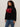 A woman with curly hair wears the Sanctuary Clothing "apres ski sweater" in black mars red (extended sizing), featuring bold red "APRÈS" lettering, paired with blue jeans against a plain, light background.