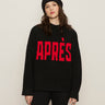 A woman with long brown hair models the Sanctuary Clothing "apres intarsia high mock logo sweater" in black/mars red, featuring bold red "APRÈS" lettering, paired with a black knit pom-pom hat and black pants against a light background.