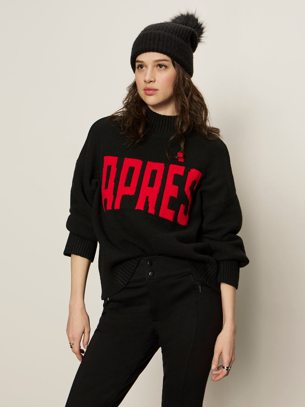 A woman stands against a plain light background wearing the Sanctuary Clothing Après Ski Sweater in black/mars red (petite sizing) and a black knit pom-pom beanie, looking off to the side in a relaxed pose.