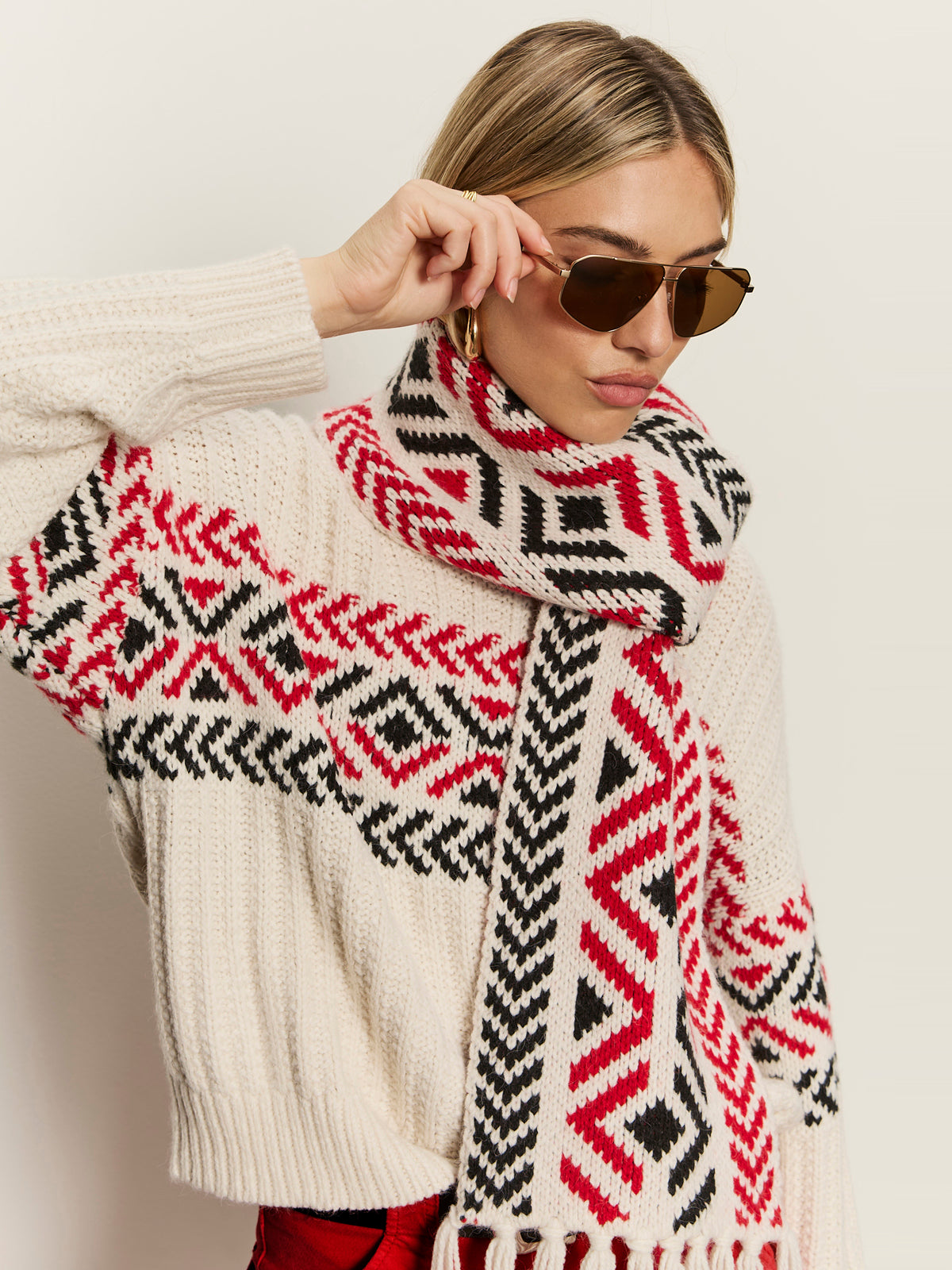 A woman poses against a plain background wearing the Sanctuary Clothing send it scarf in chalk multi, styled with aviator sunglasses and a cream knit sweater featuring red and black geometric patterns.