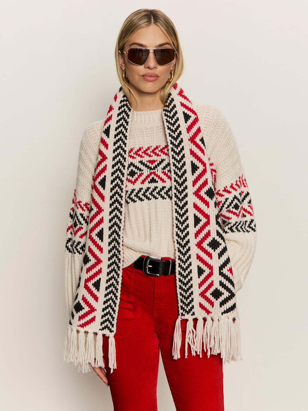 A woman wears the Sanctuary Clothing send it scarf chalk multi, paired with sunglasses, a cream sweater featuring red and black geometric patterns, and red pants against a plain background.