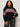 A woman with curly hair models the Sanctuary Clothing send it sweater black multi extended sizing, featuring red, white, and gray geometric patterns on the chest and sleeves, with one hand in her pocket against a plain light background.