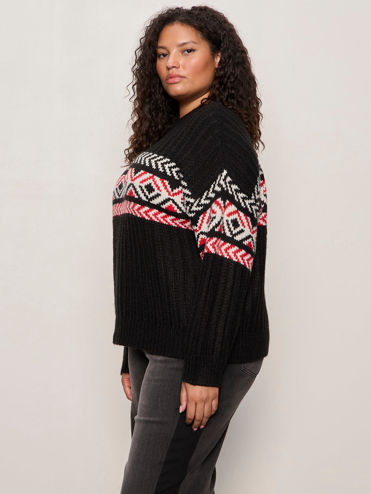 A woman with curly hair stands sideways, looking at the camera, wearing the Sanctuary Clothing send it sweater black multi extended sizing, featuring a colorful geometric pattern, against a plain light background.