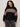 A woman with curly hair models the Sanctuary Clothing "send it sweater black multi extended sizing," a black knit featuring red, white, and beige geometric patterns. She stands against a plain light background, looking confidently at the camera.