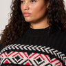 A person with long curly hair wears the "send it sweater black multi extended sizing" by Sanctuary Clothing, featuring a red and white geometric chest pattern. They look off to the side with a neutral expression.