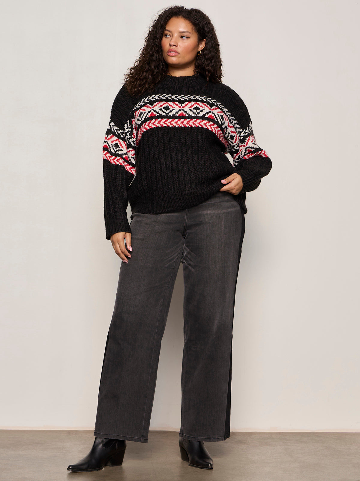 A woman wears the Sanctuary Clothing send it sweater black multi extended sizing with colorful geometric patterns, paired with dark wide-leg jeans and black ankle boots, standing against a plain light background.