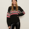 A woman models the Sanctuary Clothing "send it sweater black multi"€�a black knit with red, white, and pink geometric patterns€�and a matching beanie, posing confidently against a plain background.