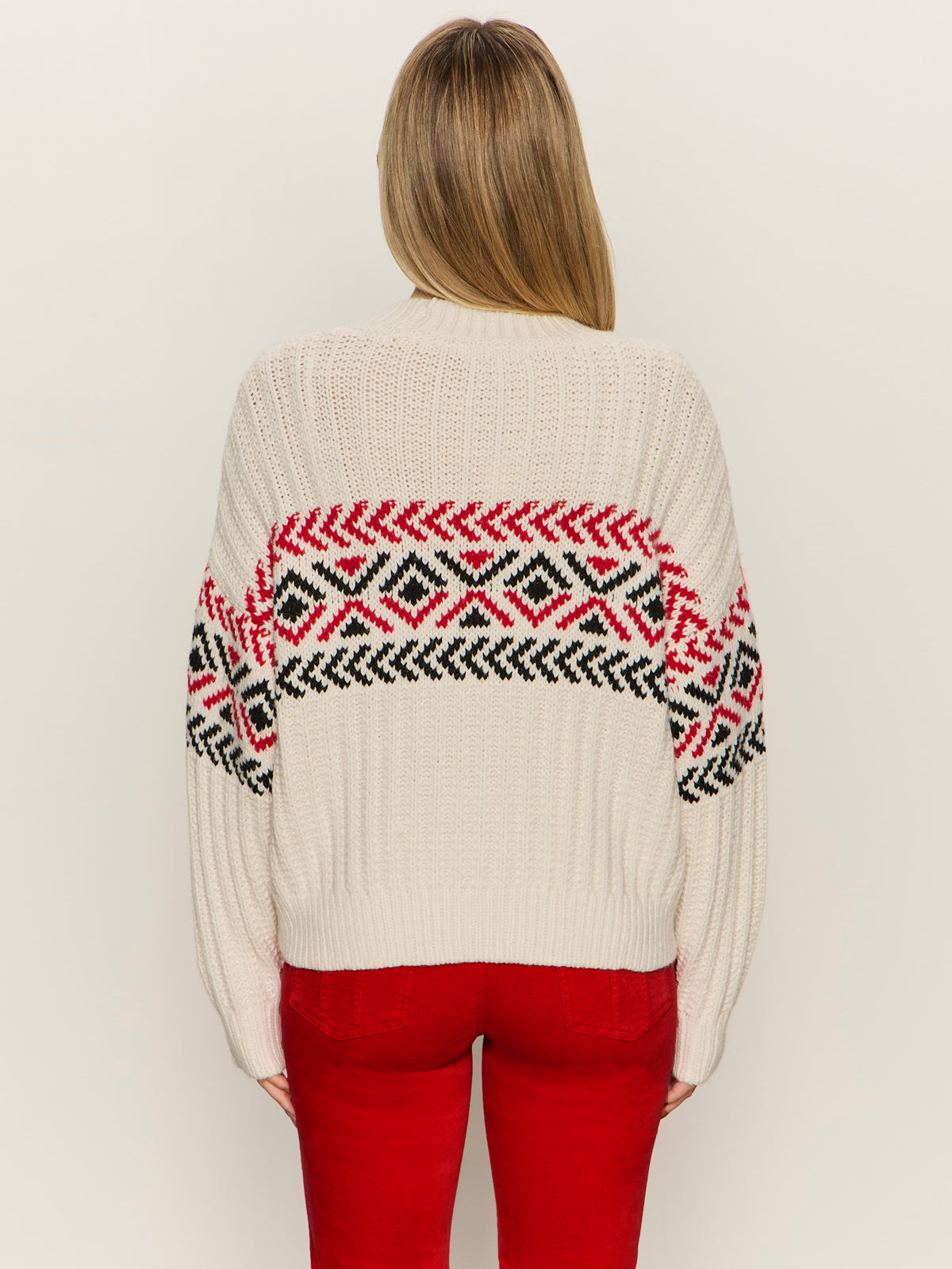 A person with long, light brown hair is seen from behind wearing the Sanctuary Clothing send it sweater in chalk multi, featuring red and black geometric patterns, paired with red pants against a plain background.