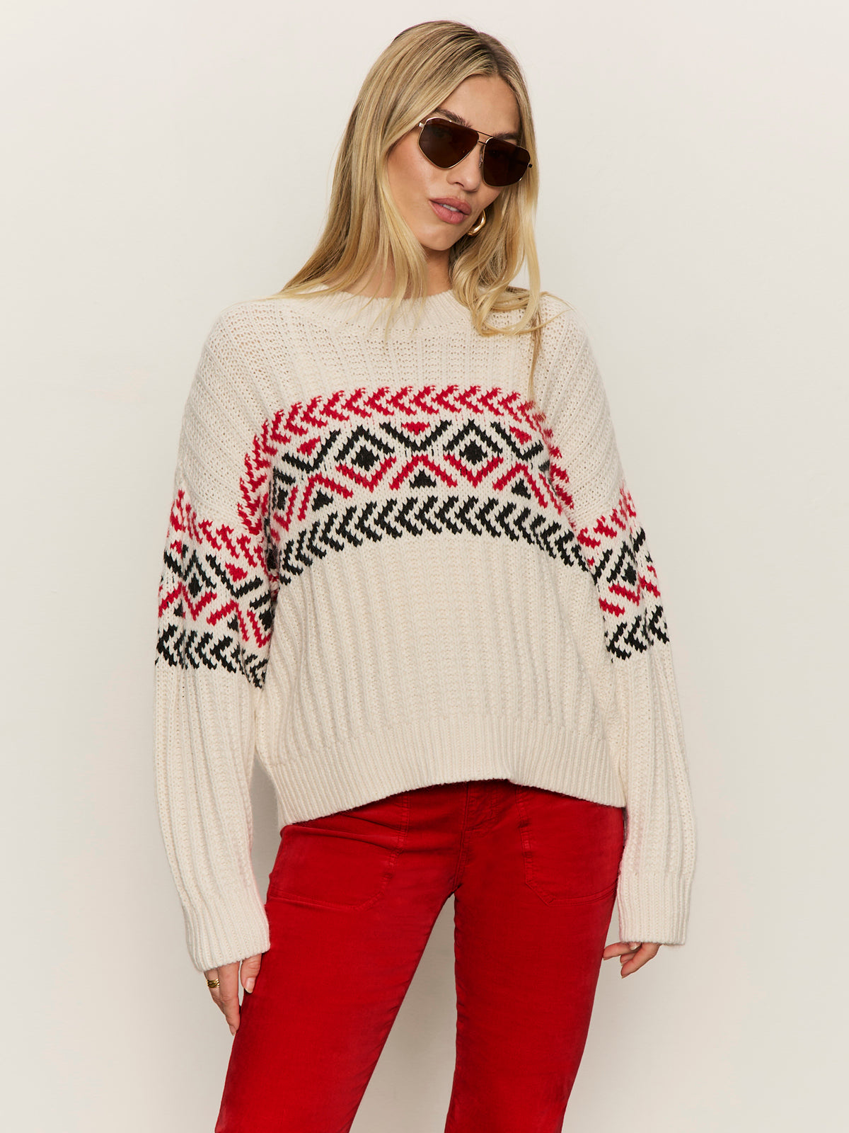A woman wears the Sanctuary Clothing send it sweater chalk multi€�cream-colored with red and black geometric patterns€�with bright red pants and sunglasses, standing against a plain light background.