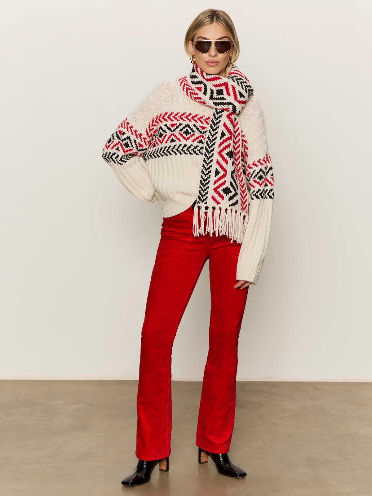 A woman models the Sanctuary Clothing "send it sweater chalk multi," featuring red and black geometric patterns, along with a matching scarf, red pants, sunglasses, and black heeled boots against a plain background.