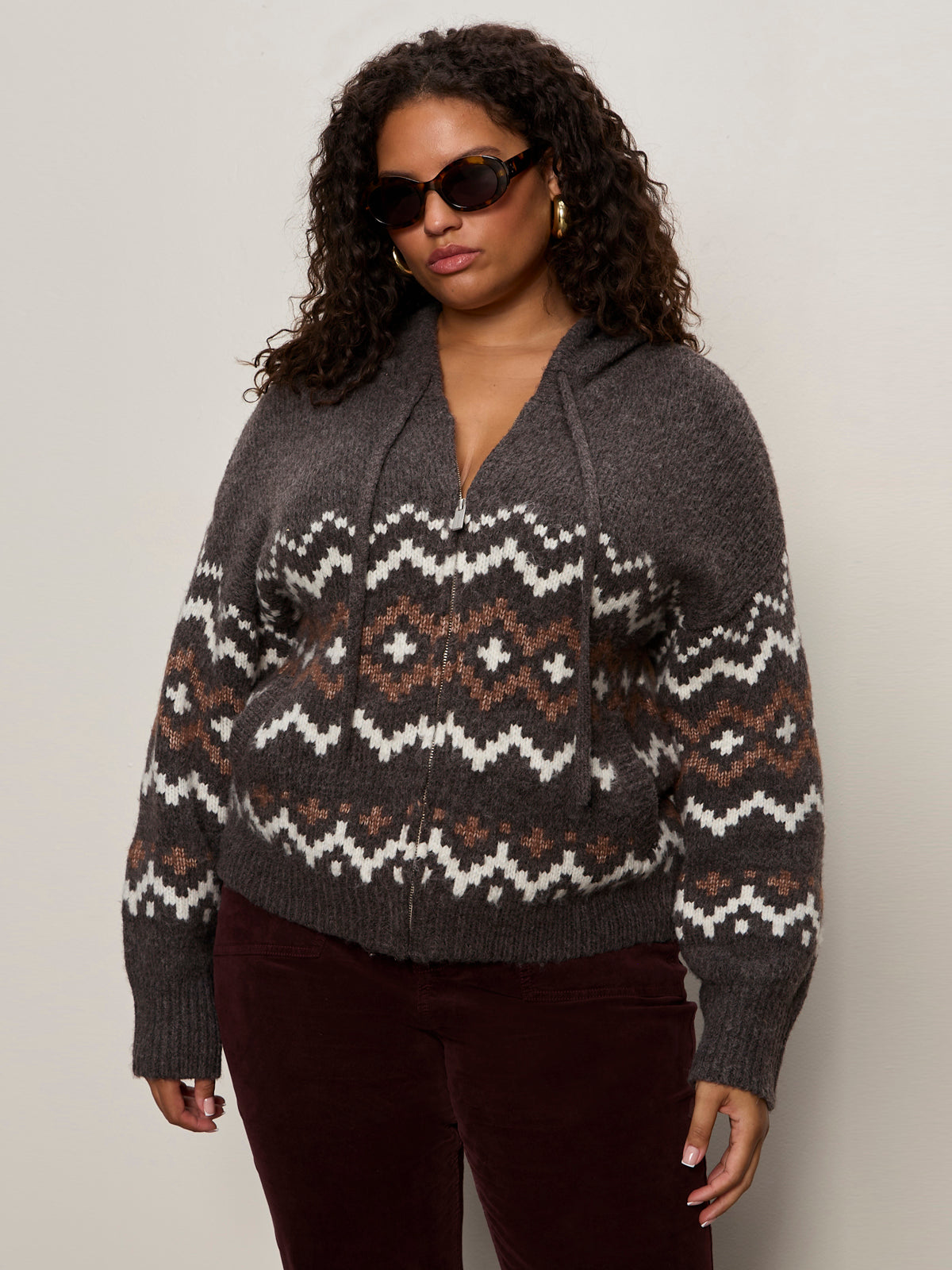 A woman with curly hair wears sunglasses, gold hoop earrings, and the Sanctuary Clothing zip up fairisle hoodie mocha fairisle extended sizing, styled with dark pants against a plain light background.
