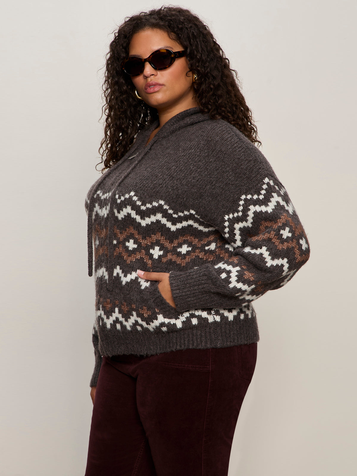 A woman with curly hair and sunglasses stands against a light background in dark pants and the Sanctuary Clothing zip up fairisle hoodie mocha fairisle extended sizing, featuring a gray and brown patterned knit, with her hand in her pocket.