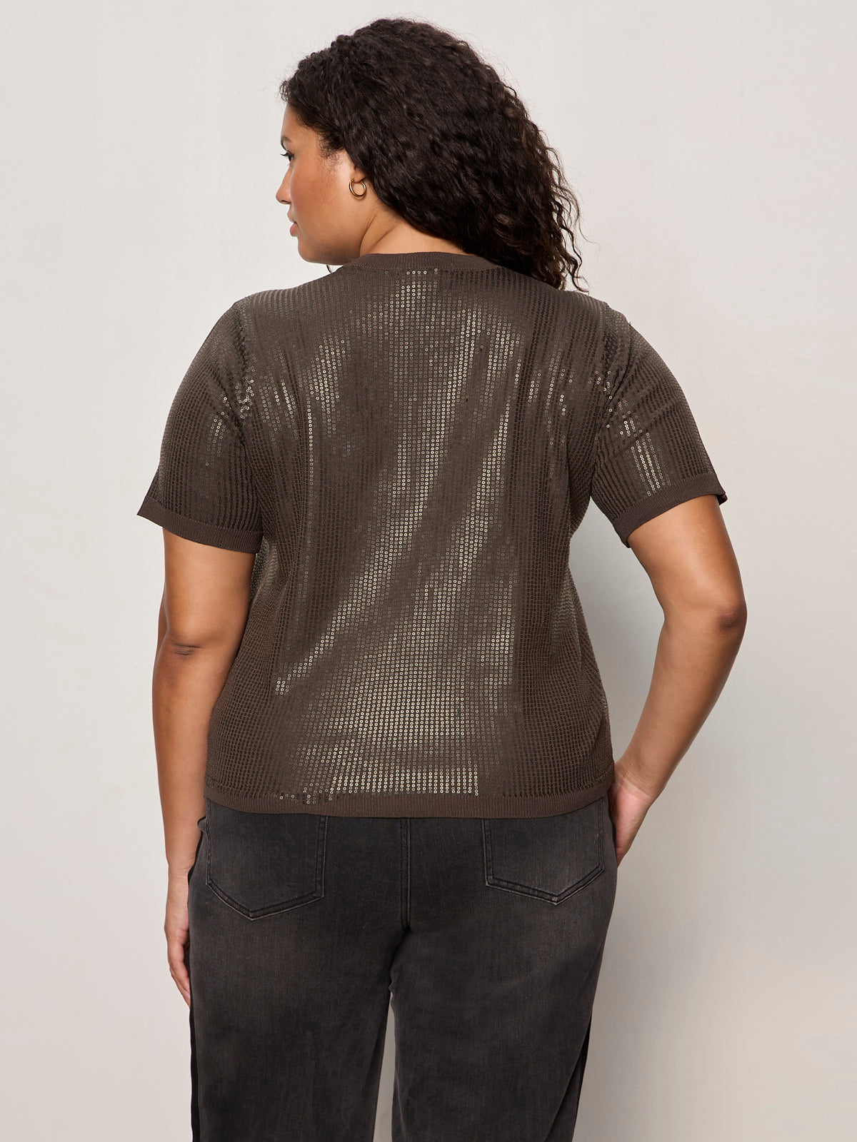 A woman with curly hair is shown from the back, wearing the Sanctuary Clothing sparkle sequin sweater ink extended sizing, standing against a plain light-colored background.