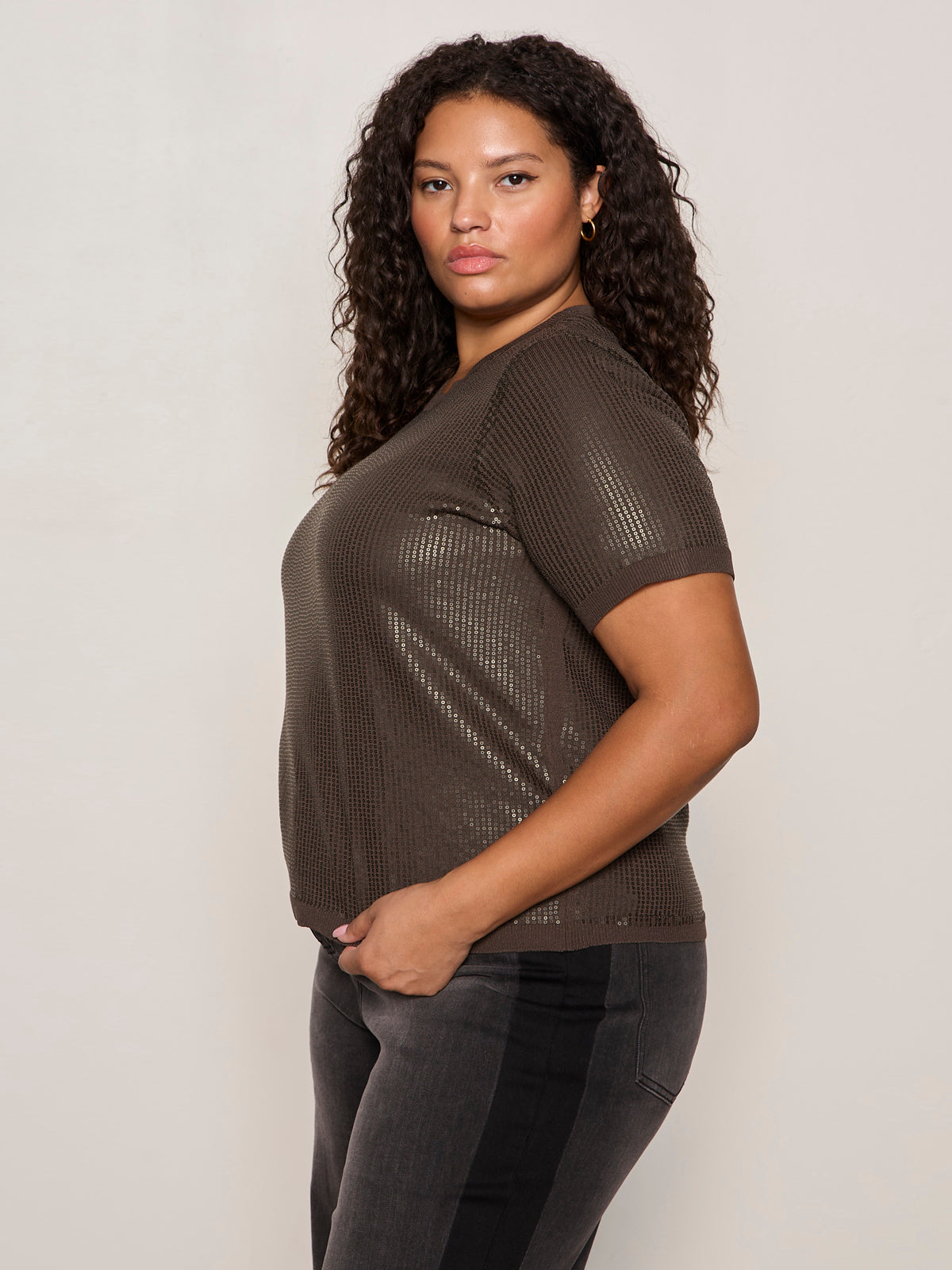 A woman with curly hair poses in profile against a plain light background, wearing the Sanctuary Clothing sparkle sequin sweater ink (extended sizing) with dark jeans, and maintains a neutral expression.