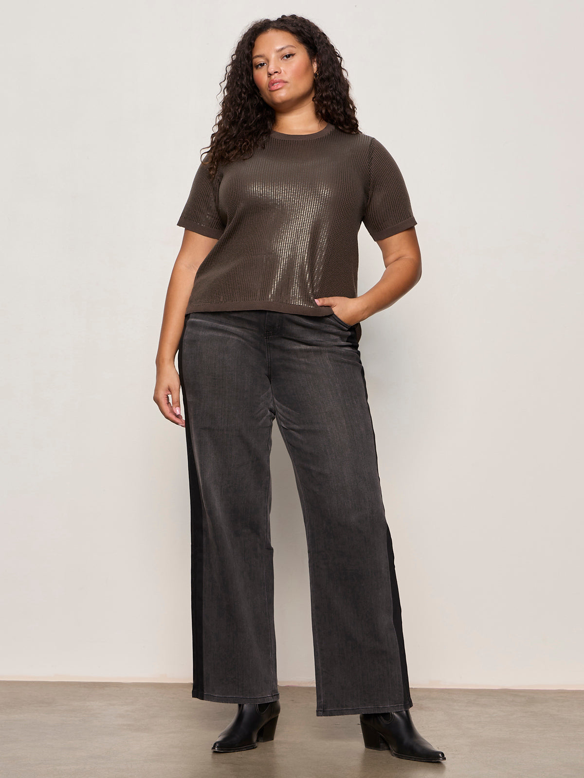 A woman with curly hair wears the Sanctuary Clothing sparkle sequin sweater ink (extended sizing), wide-leg dark gray pants, and black boots, standing confidently with one hand in her pocket against a plain light background.