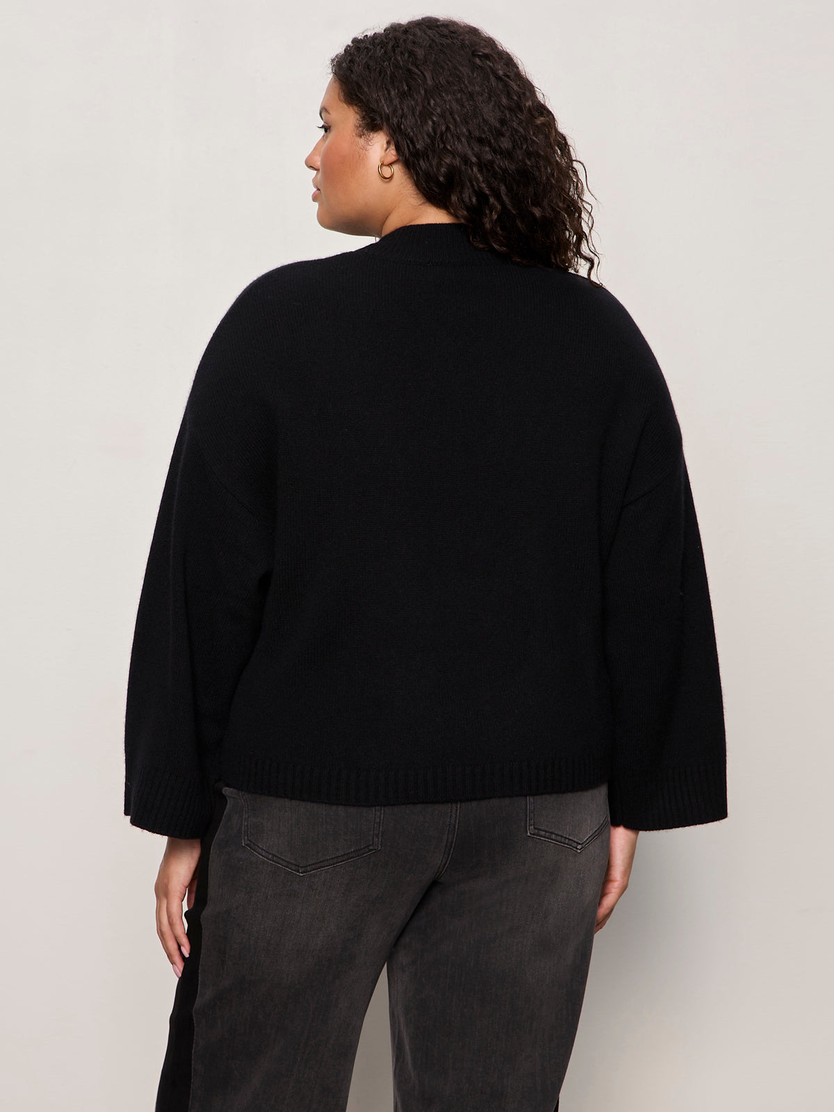 A woman with curly dark hair stands with her back to the camera, wearing the Sanctuary Clothing forever cashmere popover in black extended sizing, paired with faded black jeans against a light-colored background.