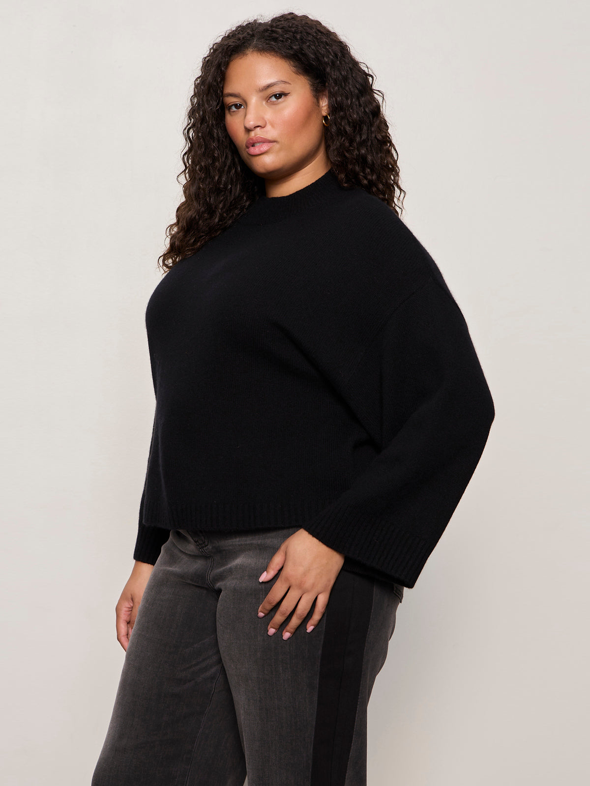 A woman with curly hair confidently models the Sanctuary Clothing forever cashmere popover in black extended sizing, paired with dark grey pants with a black stripe, standing against a light background.