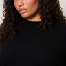 A woman with curly dark hair and natural makeup models the Sanctuary Clothing forever cashmere popover in black extended sizing, gazing confidently at the camera against a plain light background.