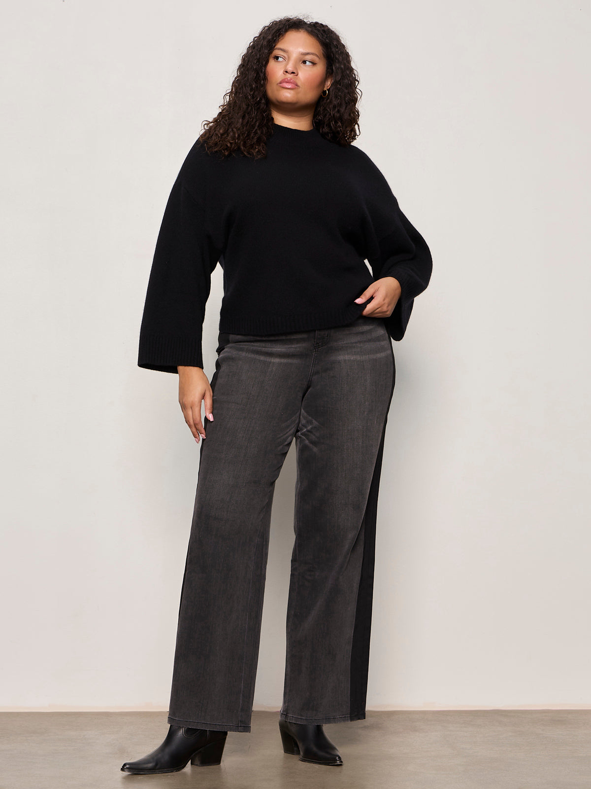 A woman with curly hair poses confidently against an off-white background wearing the Sanctuary Clothing forever cashmere popover in black (extended sizing), styled with gray wide-leg jeans and black ankle boots.