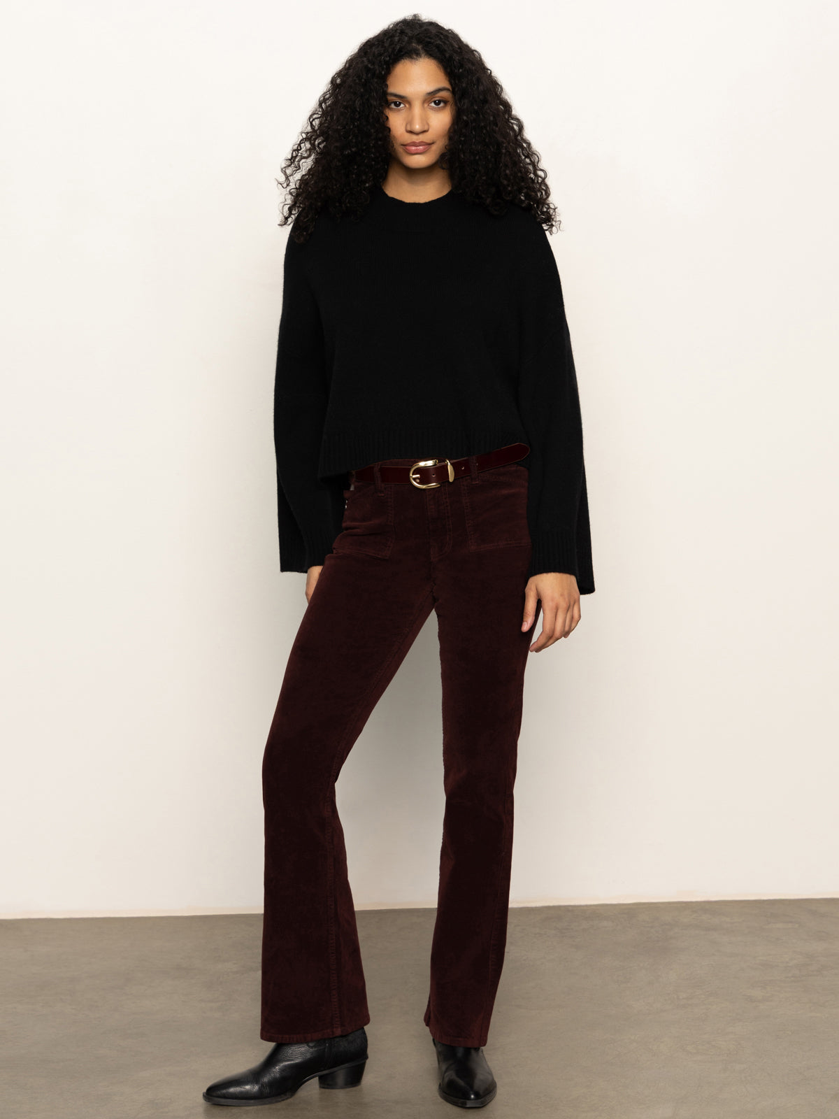 A woman with curly hair models the Sanctuary Clothing Forever Cashmere Popover in black, paired with dark red corduroy pants and black boots, standing by a light-colored wall on a concrete floor.