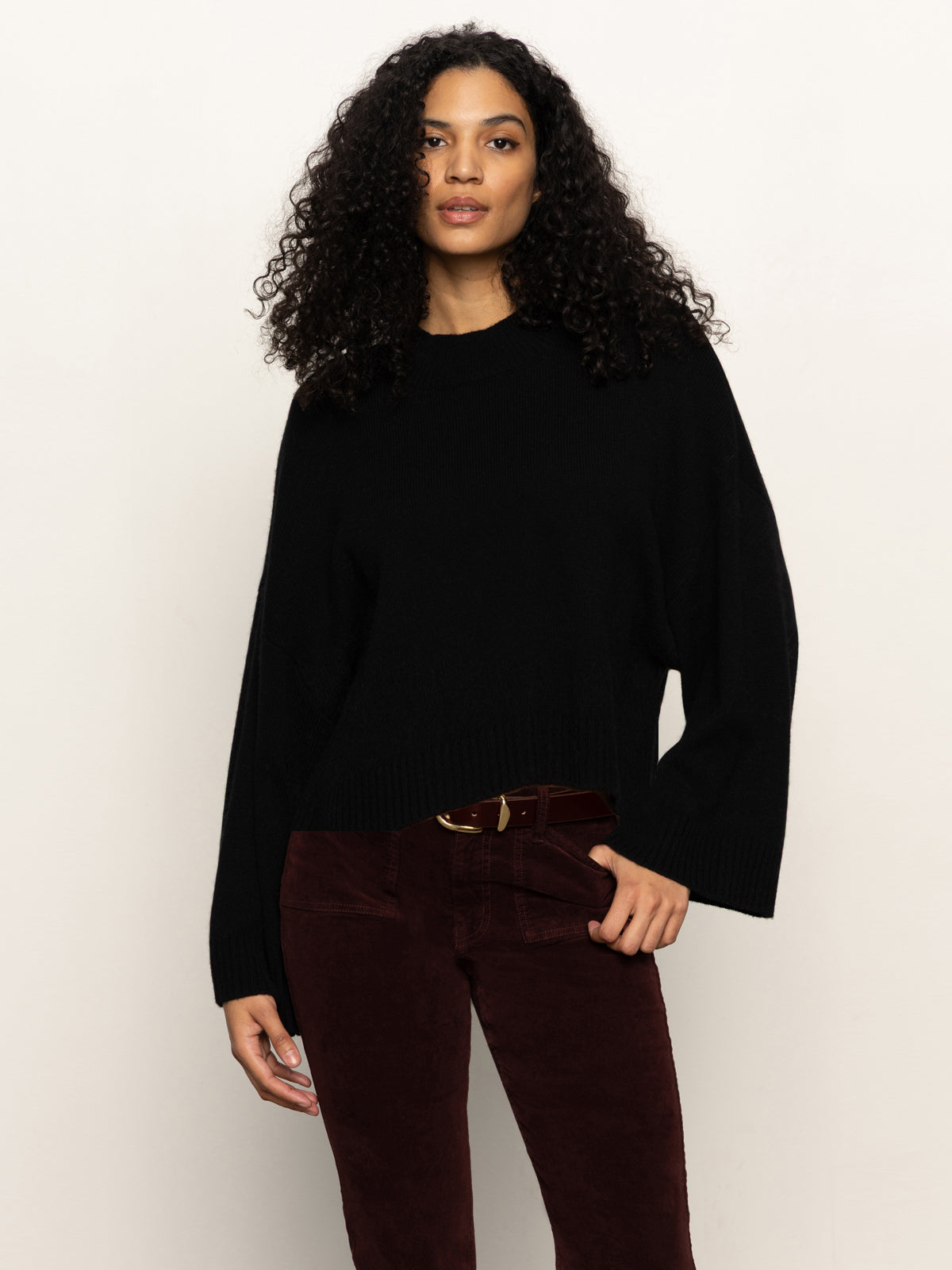 A woman with long curly hair wears the Sanctuary Clothing forever cashmere popover in black, paired with dark red corduroy pants, standing against a plain light background.