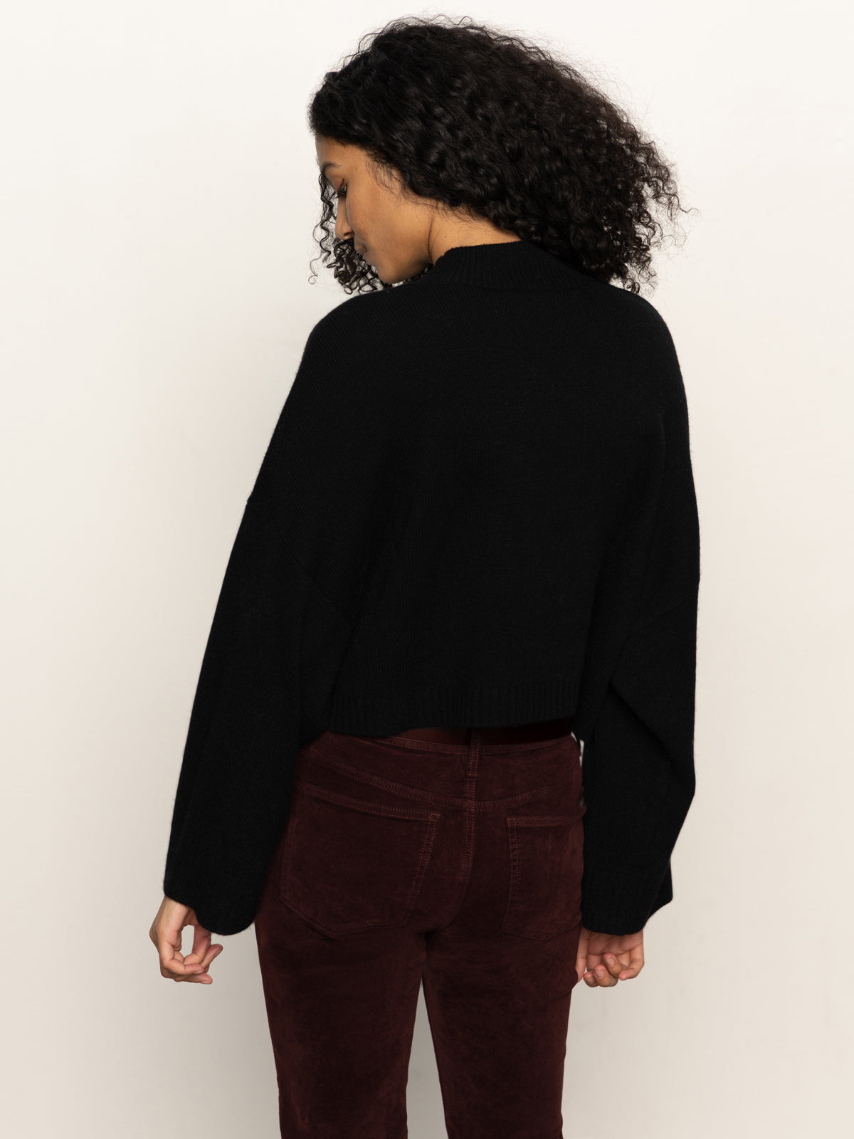 A woman with curly dark hair models the Sanctuary Clothing forever cashmere popover in black, standing with her back to the camera against a plain light background.