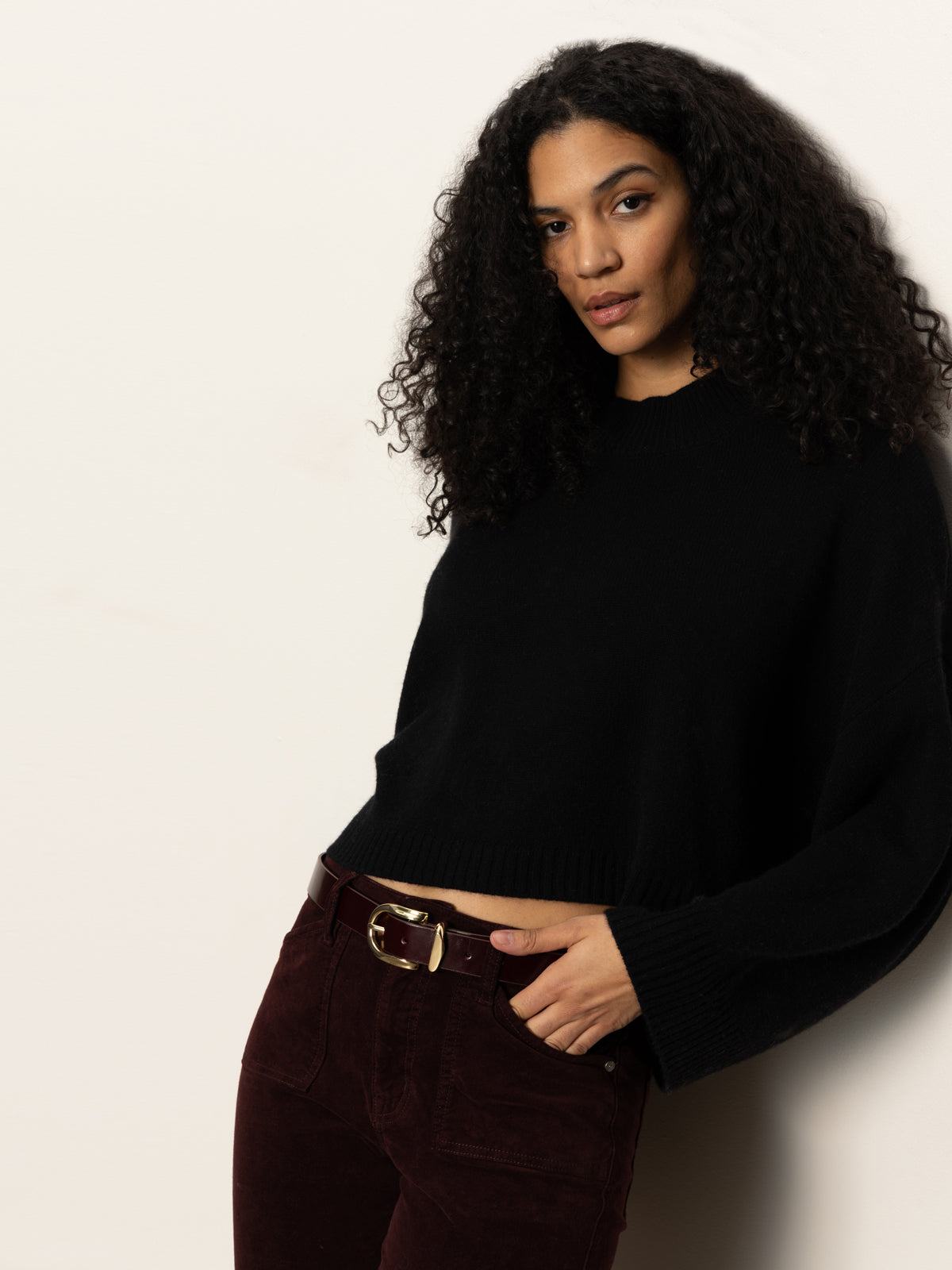 A woman with long curly hair wears the Sanctuary Clothing forever cashmere popover in black, pairing it with dark burgundy pants as she stands confidently against a light-colored wall.