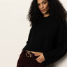 A woman with long curly hair wears the Sanctuary Clothing forever cashmere popover in black, pairing it with dark burgundy pants as she stands confidently against a light-colored wall.