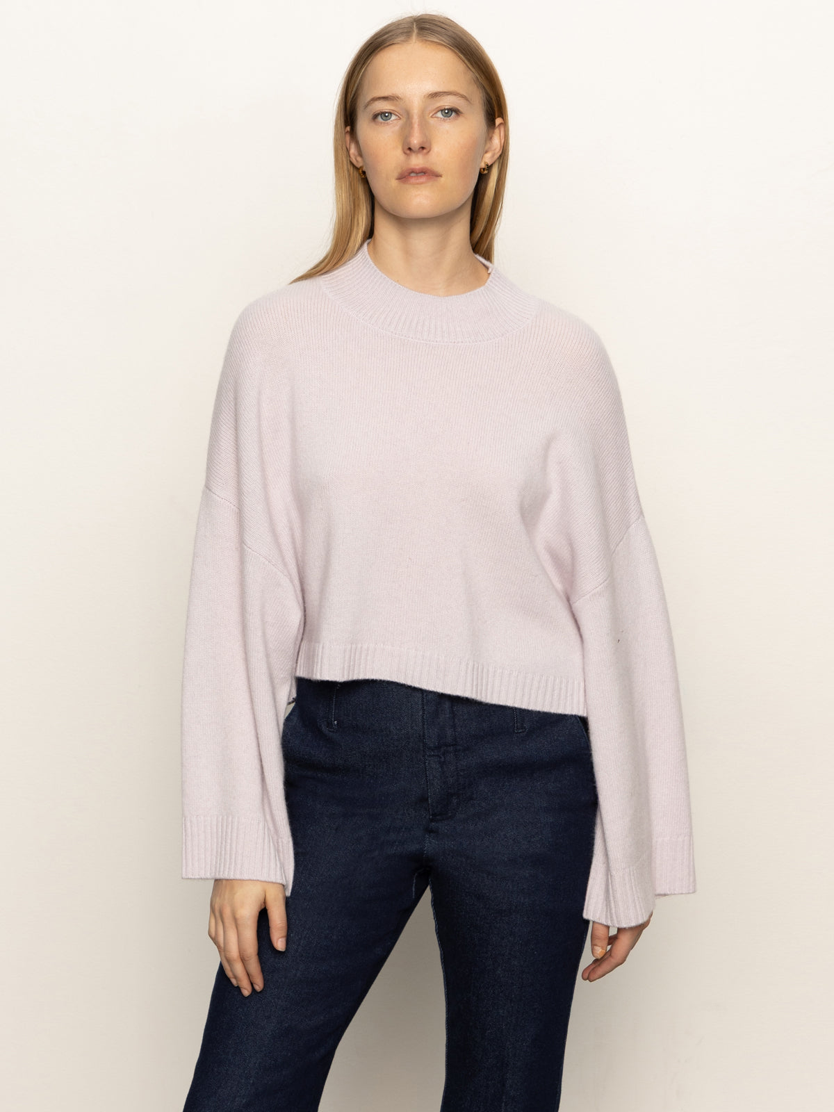 A woman with straight blonde hair wears the Sanctuary Clothing forever cashmere popover in light twilight mauve, paired with dark blue jeans, standing against an off-white background and looking at the camera.