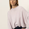 A young woman with straight, light brown hair models the Sanctuary Clothing forever cashmere popover in light twilight mauve, paired with dark pants. She poses against a plain, light background with a neutral expression, gazing slightly away.
