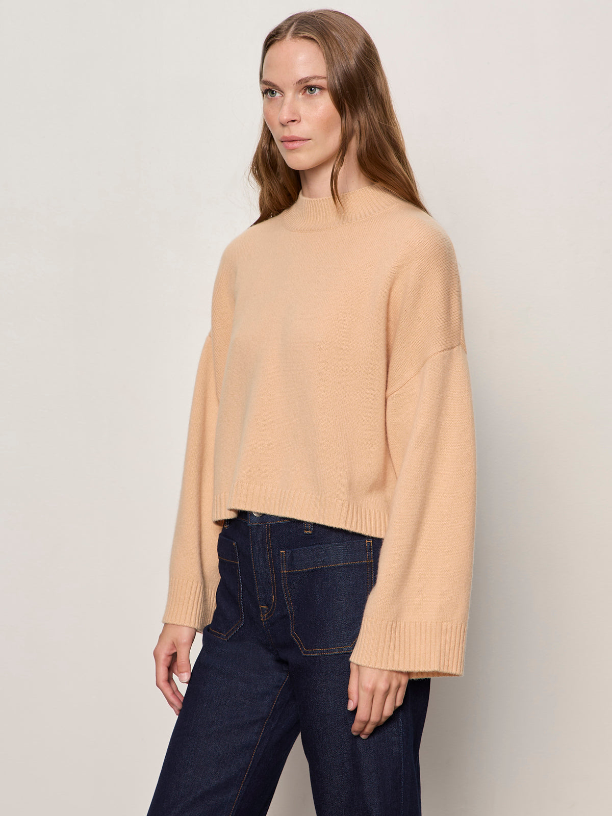A woman with long, straight brown hair wears the Sanctuary Clothing forever cashmere popover in toasted beige, paired with dark blue high-waisted jeans, standing against a plain light background.