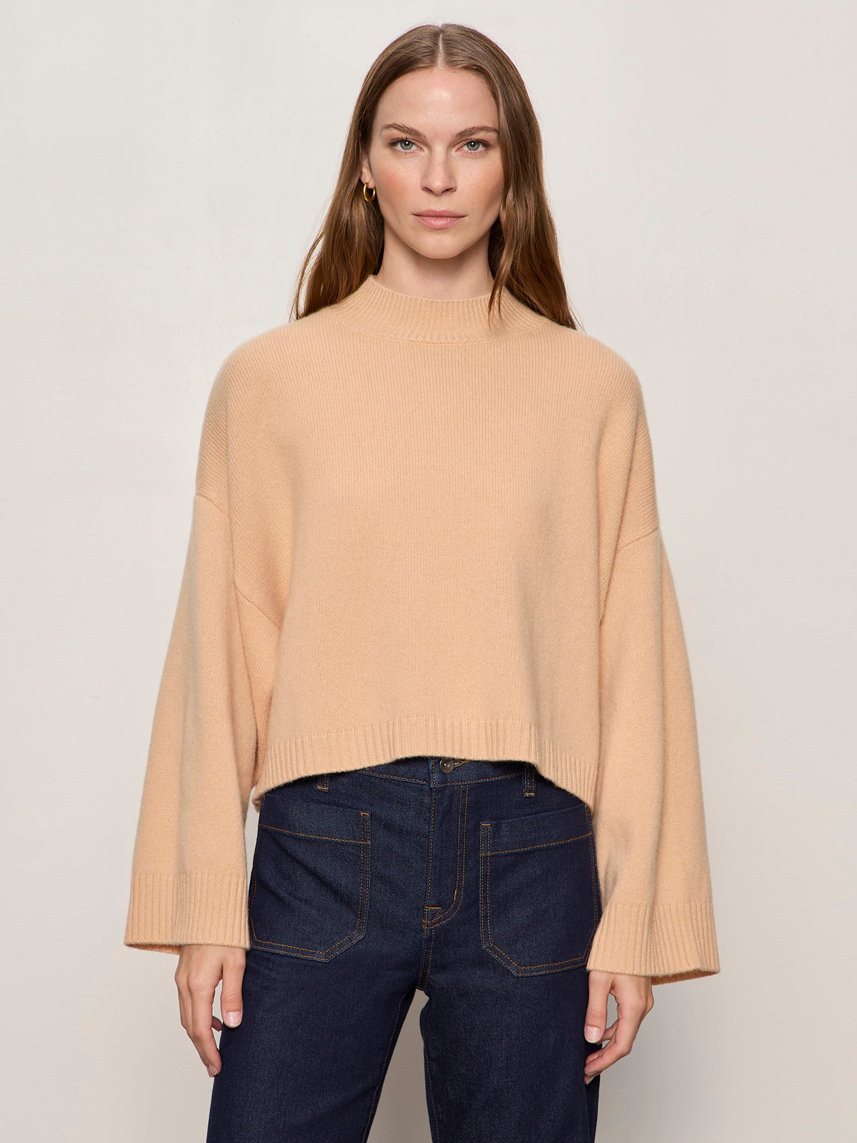 A woman with long brown hair wears the Sanctuary Clothing Forever Cashmere Popover in toasted beige and dark blue high-waisted jeans, standing against a plain, light background.