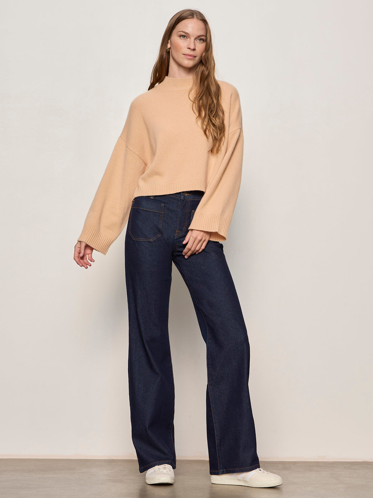 A woman with long, wavy hair stands against a light wall, wearing the Sanctuary Clothing forever cashmere popover in toasted beige, paired with dark blue wide-leg jeans and white sneakers. She exudes a relaxed, confident vibe.