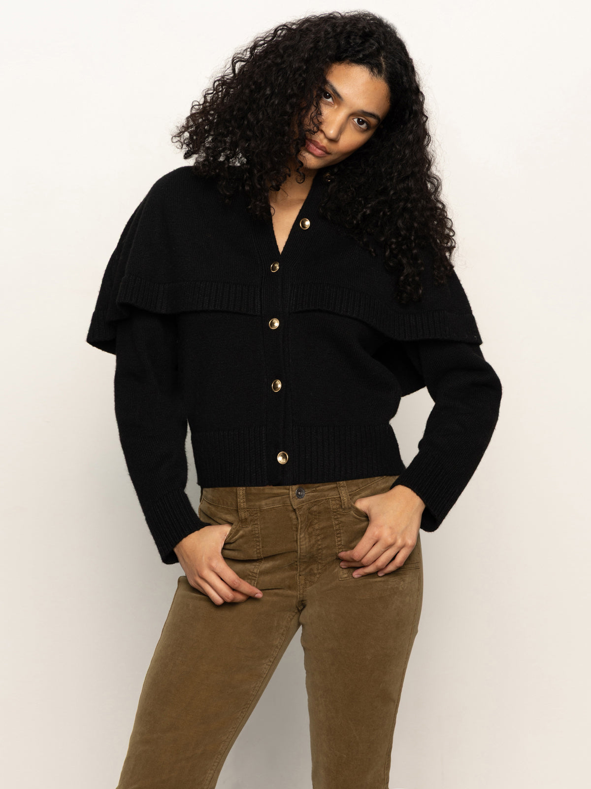 A woman with curly dark hair models the Sanctuary Clothing cape cardi black sweater, featuring a wide collar, paired with brown pants against a plain light background.