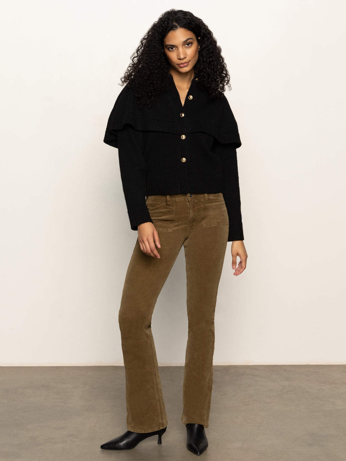 A woman with curly hair stands against a light background, wearing the Sanctuary Clothing cape cardi black sweater, brown corduroy pants, and black boots. She poses relaxedly with one arm by her side and the other slightly bent.