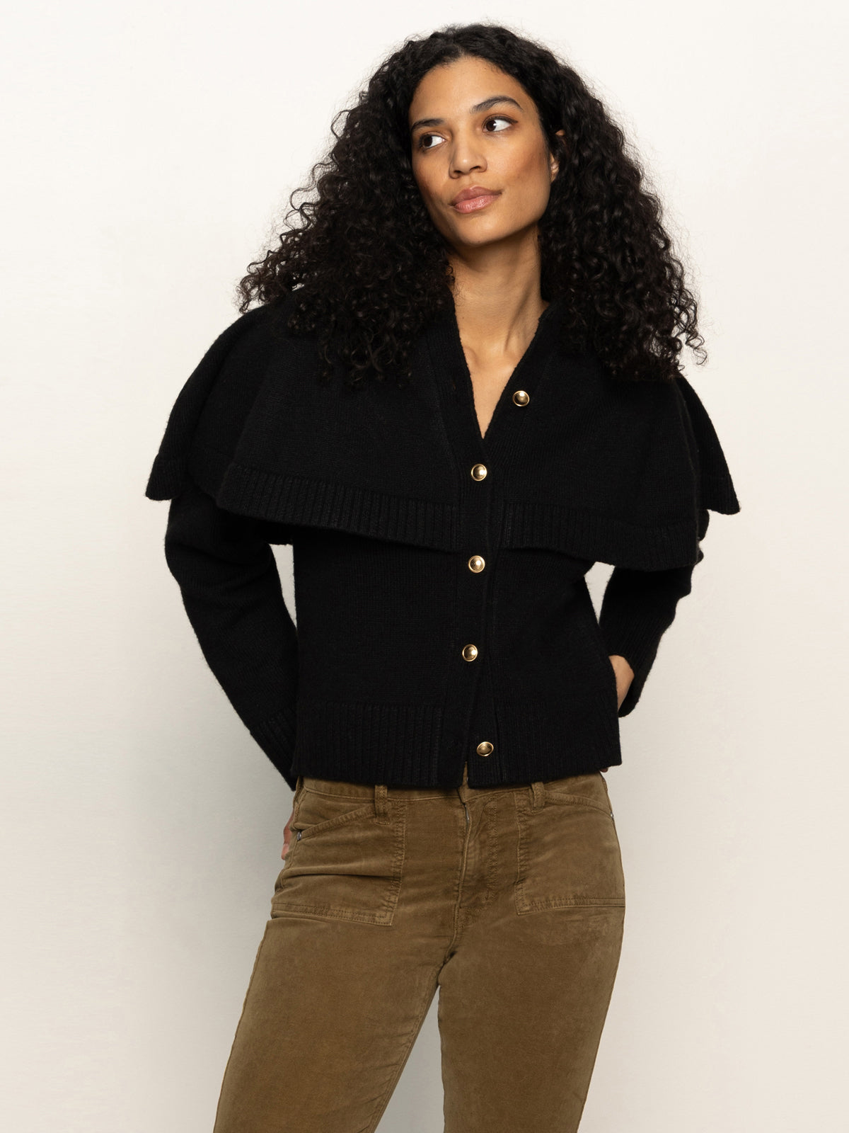 A woman with curly hair wears the Cape Cardi Black sweater by Sanctuary Clothing and brown pants. She stands against a light background with her hands in her pockets, gazing slightly to the side.
