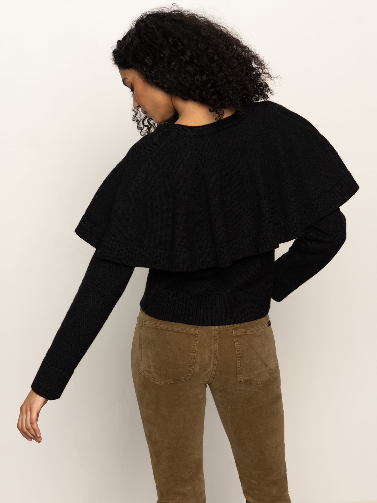 A person with curly hair models Sanctuary Clothing's cape cardi black, styled with brown pants, standing with their back to the camera against a plain light background.