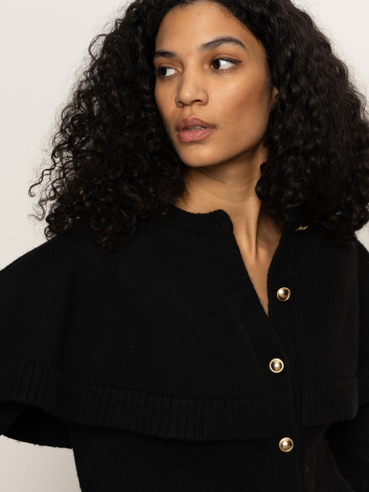 A woman with curly dark hair wears the Sanctuary Clothing cape cardi in black and looks to the side against a plain background.