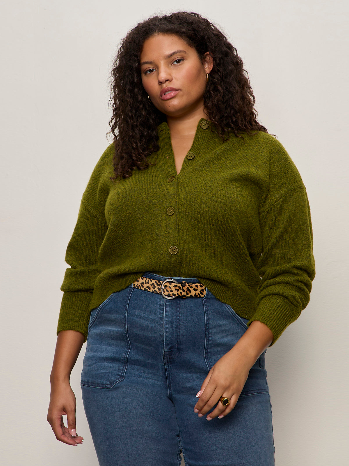 A woman with curly hair wears a green button-up sweater tucked into high-waisted blue jeans, styled with the Sanctuary Clothing riot leopard belt leopard, standing against a plain light background.