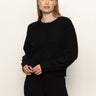 A woman with straight, light brown hair is wearing the Sanctuary Clothing must have cardi black sweater and black pants, standing against an off-white background with a neutral expression.