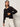 A woman wears the Sanctuary Clothing for keeps pointelle cardigan in black over a black top and white pants, standing against a plain light background and looking to the side with one hand on her waist.