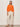 A woman with straight dark hair wears the Sanctuary Clothing for keeps pointelle cardigan in hibiscus orange, paired with off-white pants and beige slide sandals, standing against a plain light wall with minimal jewelry and a neutral expression.
