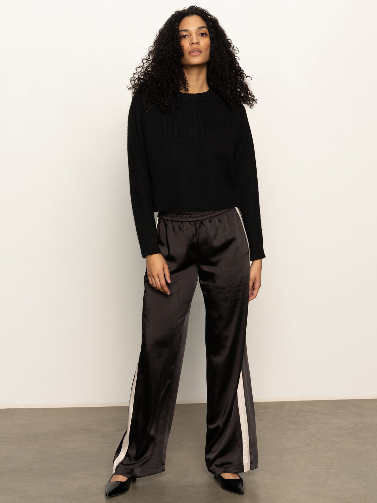 A woman with curly hair wearing the Sanctuary Clothing zip it sweater in black stands against a plain light-colored wall on a concrete floor, paired with loose shiny black pants with white side stripes.