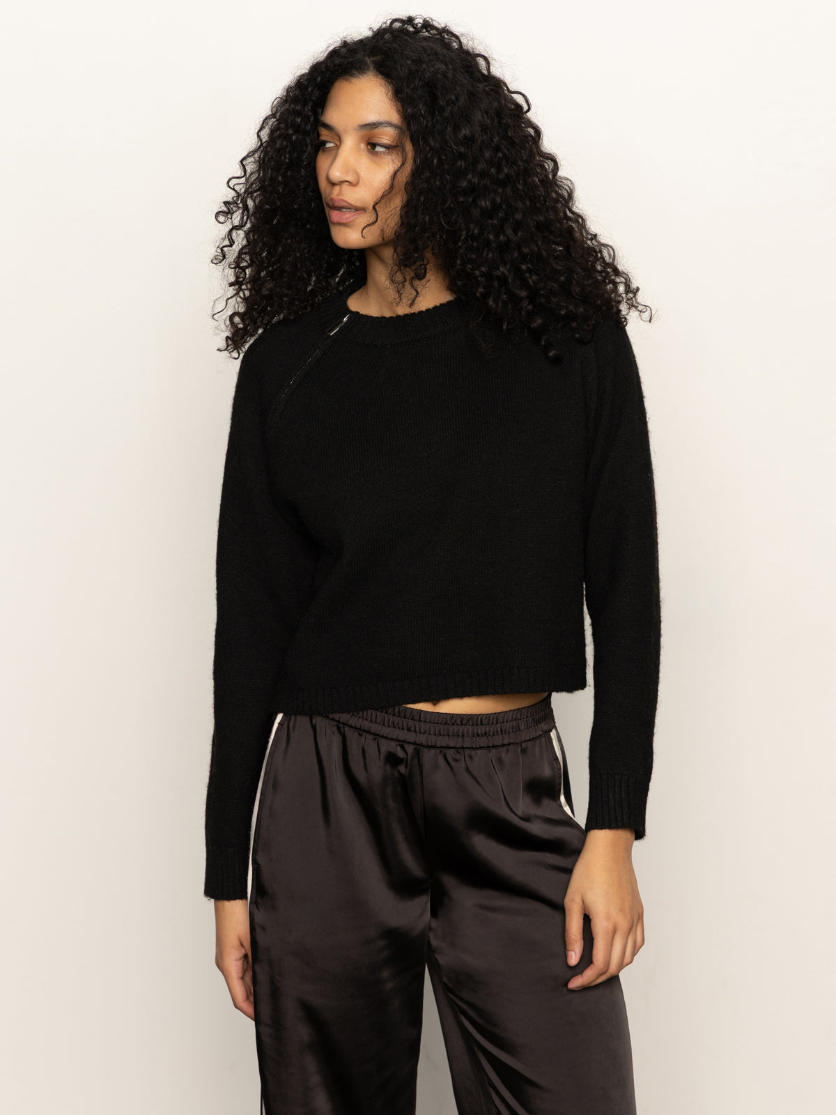 A woman with long, curly hair models the Sanctuary Clothing "zip it sweater black" against a plain light background, pairing it with silky dark pants featuring white side stripes and looking slightly to the side with a relaxed expression.